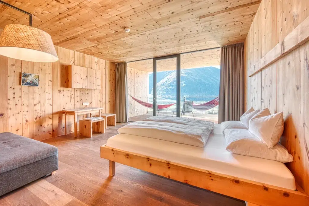Schlafzimmer mit Holzvertäfelung, ausgestattet mit Bett, Schreibtisch und Sofa; vom Balkon aus hat man einen Blick auf die Berge, und draußen stehen Hängematten.