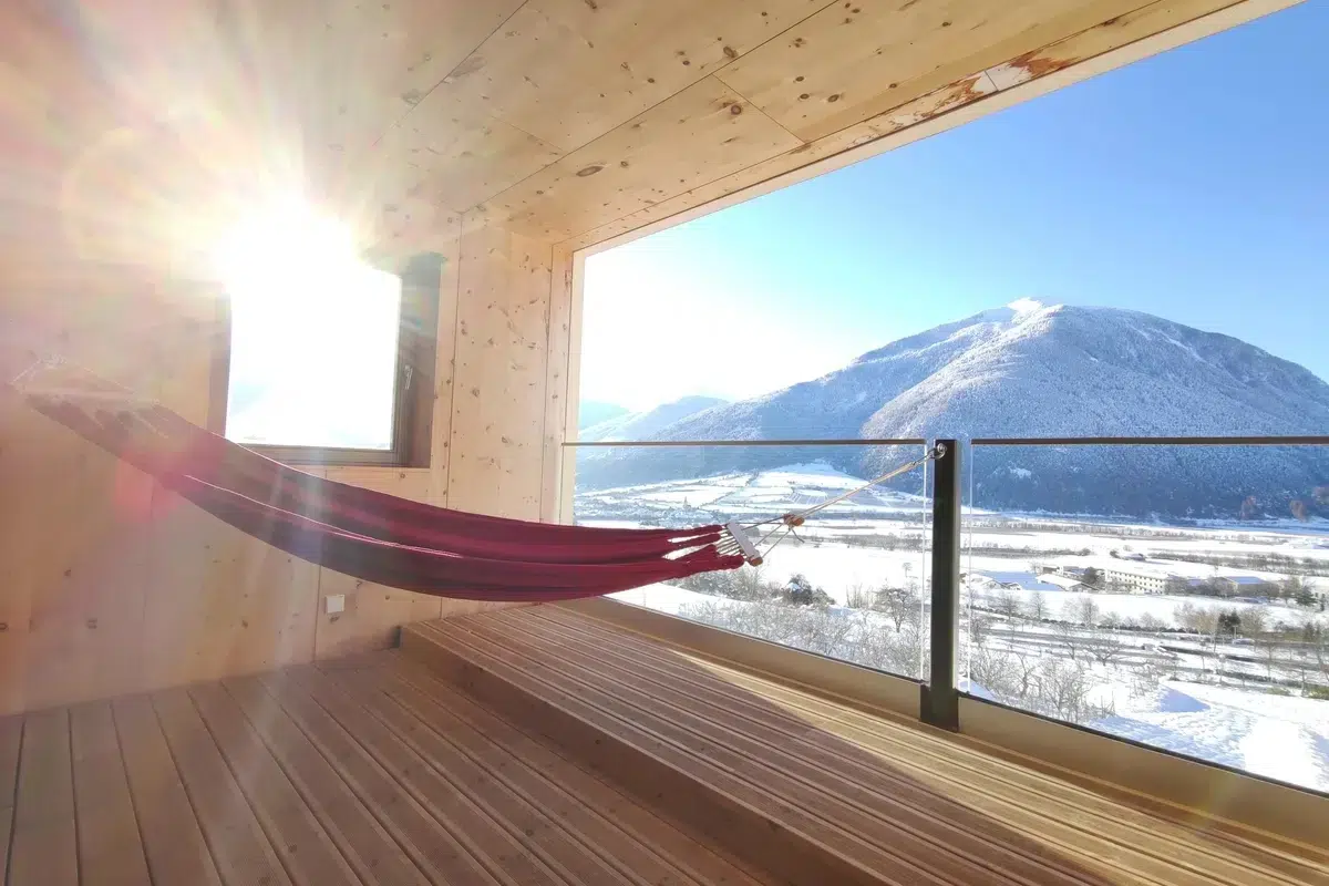 A hammock on a wooden balcony overlooks snowy mountains in bright sunlight.